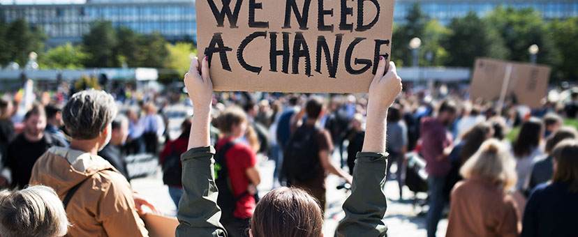 Woman at protest holds sign that reads “We Need Change”